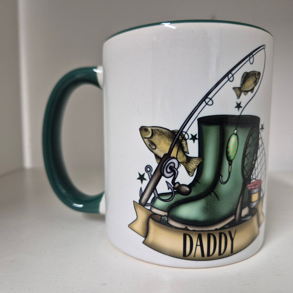 White ceramic mug with hunter‑green handle featuring fishing boots, rod with leaping fish, landing net, bait bucket and “Daddy” banner illustration